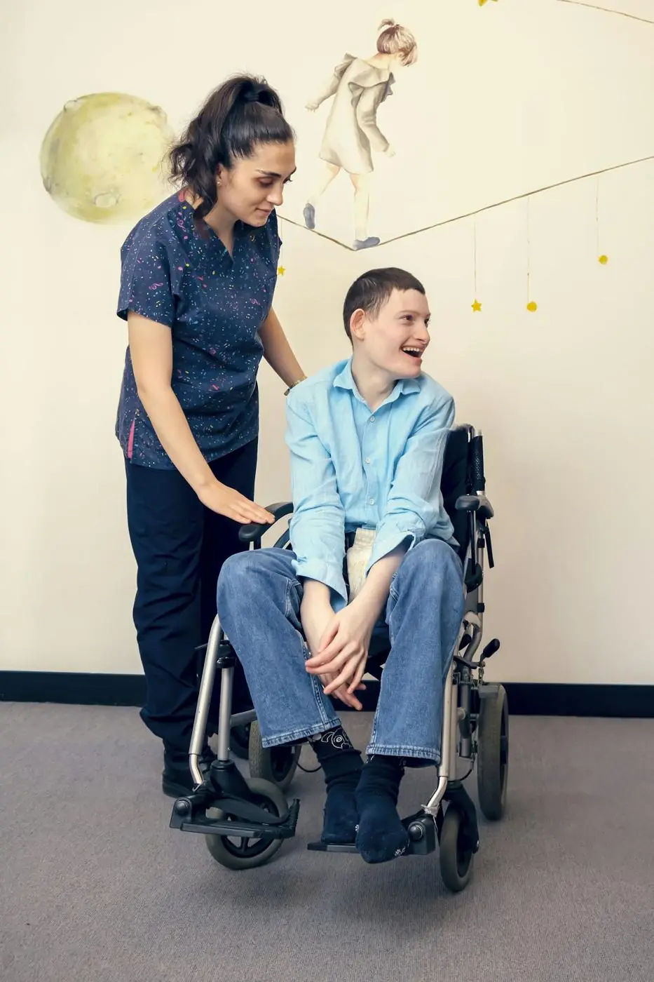 A support worker standing beside a young man using a wheelchair, both smiling